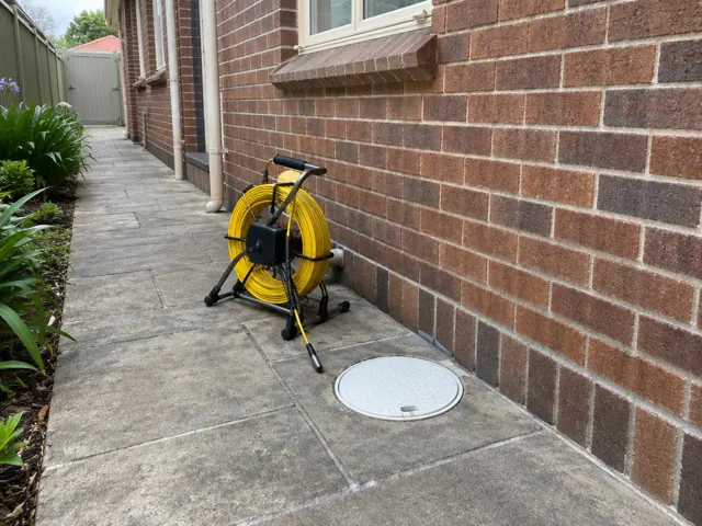 Capped drain inspection cover beside a coiled camera reel on a paved side path by a brick wall in Malabar