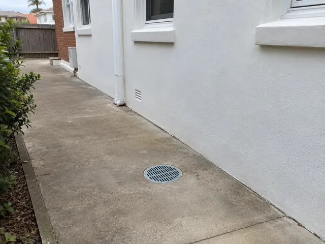 Outdoor concrete gully grate beside a rendered house wall and side path in Maroubra