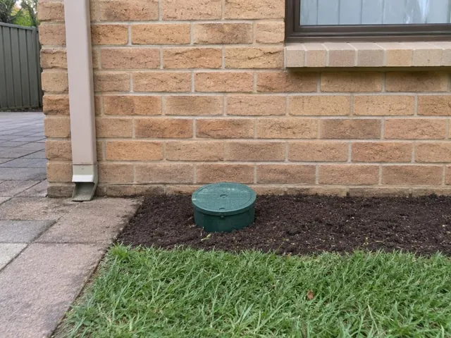 Green capped sewer opening in a garden bed beside a brick house wall in Matraville